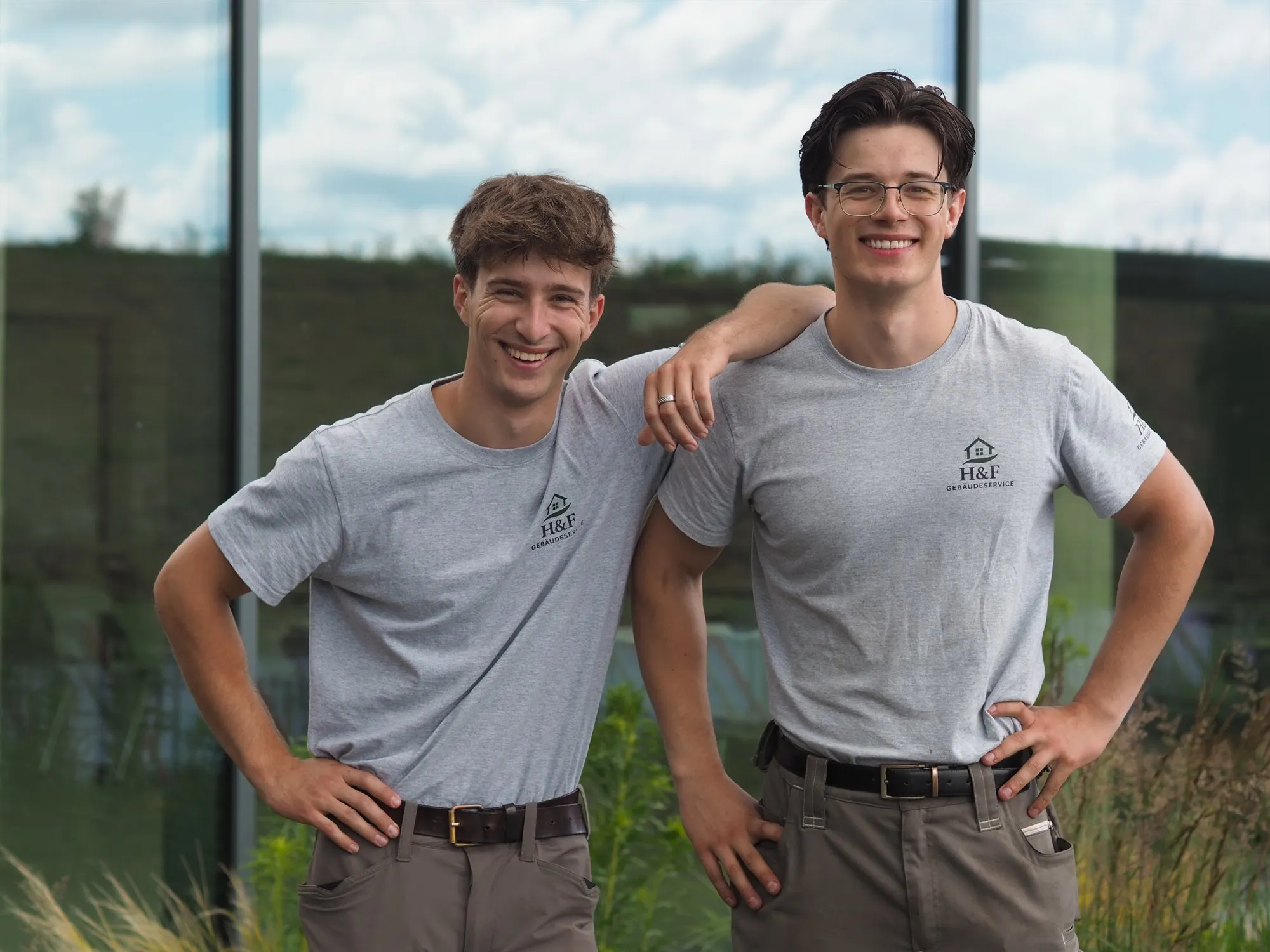 Die Gründer von H&F Gebäudeservice, Max Haustedt und Max Förster, in grauen H&F-Shirts vor einer Glasfassade in Wien